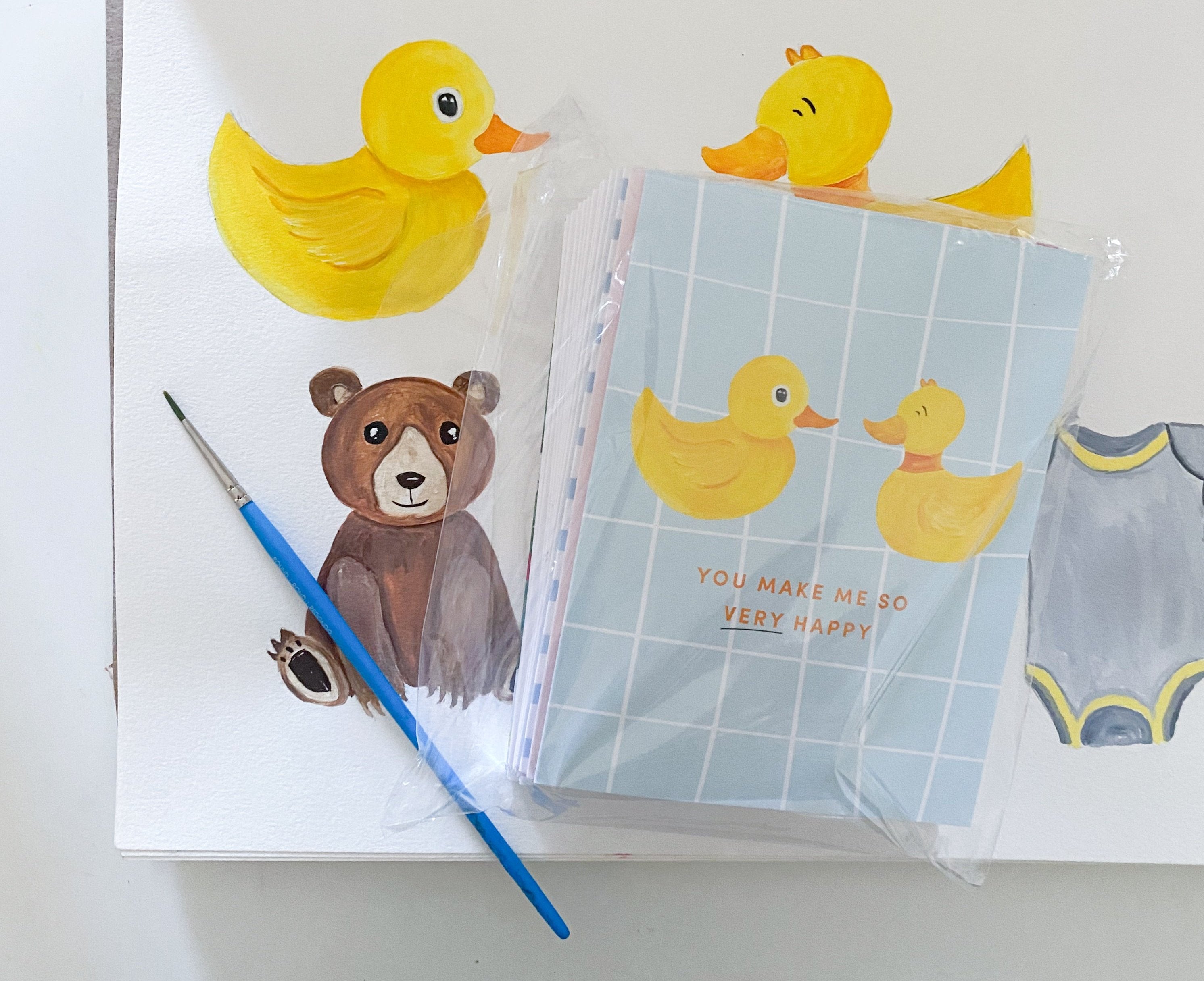 Painting of two yellow ducks, a teddy bear and baby onesie. A greeting card with two ducks sits on top of the painting. It reads You Make Me So Very Happy.